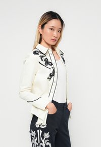 White button-up shirt with black floral embroidery and trim, paired with dark pants featuring similar white floral detailing.