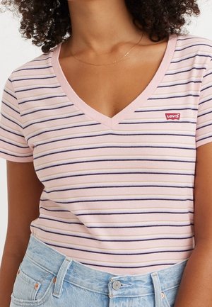 Frau trägt ein pink- und marineblau gestreiftes V-Ausschnitt-Levi's-T-Shirt, das in hellblaue Jeans gesteckt ist, dazu eine dünne goldene Halskette.