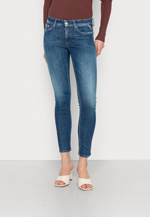 Jean slim bleu avec légère décoloration, porté avec un haut marron à manches longues côtelées et des sandales blanches à talons ouverts.