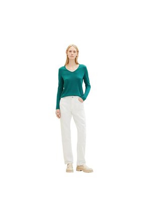 Femme se tenant debout, portant un haut en V à manches longues teal, un pantalon blanc taille haute et des chaussures à semelle épaisse beige, avec une main dans la poche.