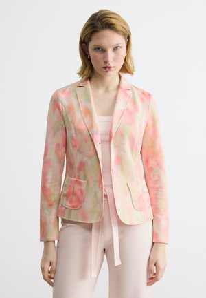 Femme portant un blazer à motifs rose pastel et vert par-dessus un haut et un pantalon rose clair, debout devant un fond clair uni.