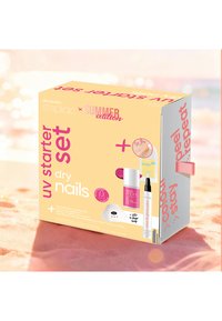 Kit de démarrage UV pour ongles secs avec vernis rose, lampe LED 5 doigts, stylo de soin et brillance, et sachet de crème pour les mains à l'urée sur un fond de plage.