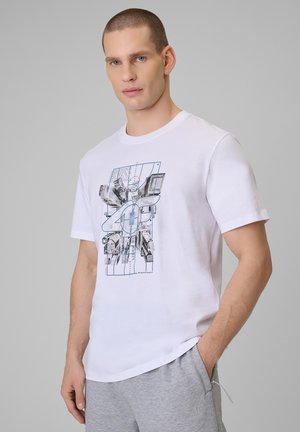 Mężczyzna w białym t-shircie z abstrakcyjnym wzorem i jasnoszarych spodniach dresowych, stoi z jedną ręką w kieszeni na jednolitym tle.