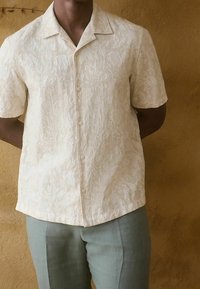 Camisa de manga corta beige con un patrón texturizado de hojas, botones en la parte delantera, combinada con pantalones cortos de color verde claro.