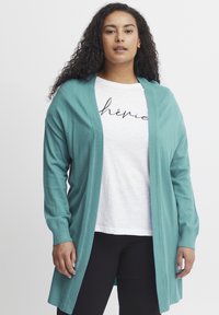 Turkos kofta med öppen front, långa ärmar och mjuk struktur, lager på lager över en vit T-shirt med svart skripttext.