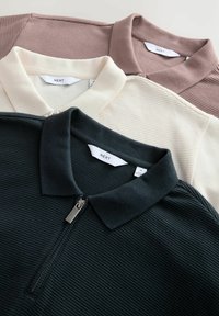 Tres camisas polo en verde oscuro, crema y malva claro. Las características incluyen textura acanalada, cuello clásico y un frontal con cremallera en la camiseta verde oscuro.
