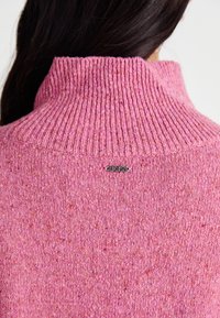 Pull en tricot rose avec un col côtelé haut, un tissu texturé avec des speckles, et un détail de logo argenté à l'arrière du cou.