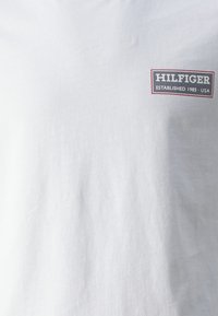 Bílý bavlněný tričko s obdélníkovým logem a nápisem "HILFIGER" a "ESTABLISHED 1985 - USA" v stylizovaném písmu.