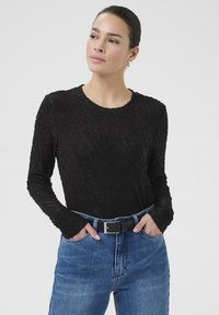 Sort langærmet top med tekstureret mønster, rund halsudskæring og tætsiddende ærmer, parret med blå højdenede jeans og et sort bælte.