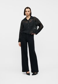Femme aux cheveux courts et foncés portant une chemise noire transparente boutonnée, un pantalon noir taille haute à jambes larges, et des talons noirs pointus, debout avec une main sur la taille.