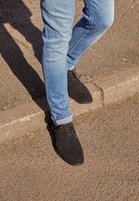 Personne portant des chaussures noires et un jean bleu descend d’un trottoir sur une surface pavée au soleil.