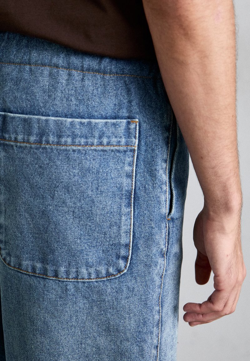 Jeans in denim di colore azzurro chiaro con cuciture arancioni a contrasto, dotati di una tasca posteriore e design a gamba dritta. Texture morbida e vestibilità casual.