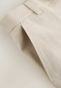 Gros plan sur la poche d'un pantalon en tissu beige avec des coutures visibles et une passants de ceinture, sur un fond clair.