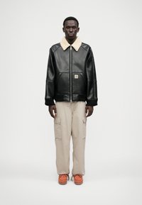 Czarna skórzana kurtka z kremowym kołnierzem z koźlej skóry, w połączeniu z beżowymi spodniami cargo i pomarańczowym obuwiem. Z przodu zamek błyskawiczny i kieszenie.