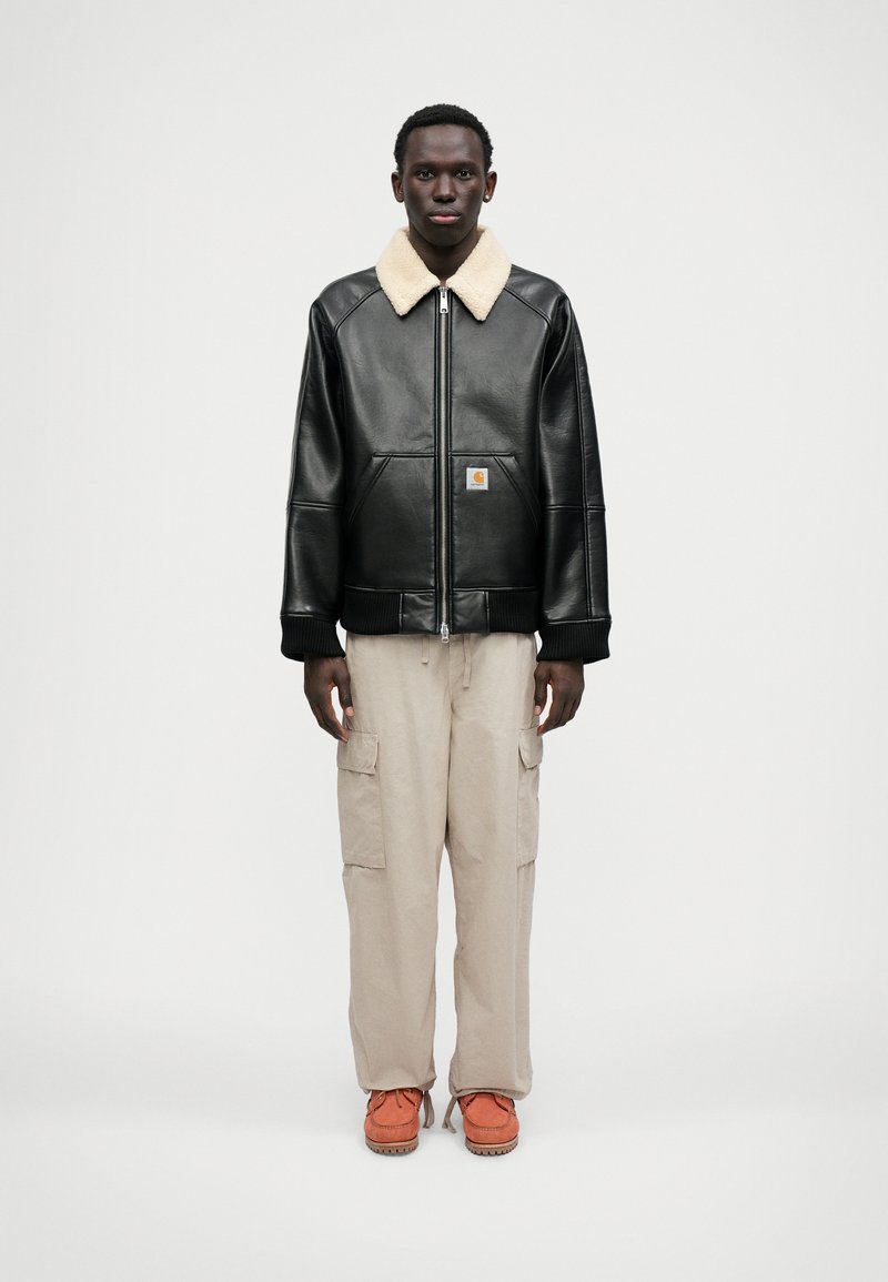 Schwarze Lederjacke mit einem cremefarbenen Lammfellkragen, kombiniert mit beigen Cargo-Hosen und orangefarbener Fußbekleidung. Mit einem Frontreißverschluss und Taschen.