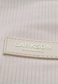 Homokszínű textil táska, amely texturált hálós mintával rendelkezik. Egy téglalap alakú világos homokszínű címke található rajta, amelyen a "LARKSON HAMBURG" felirat dombornyomott szöveggel olvasható.