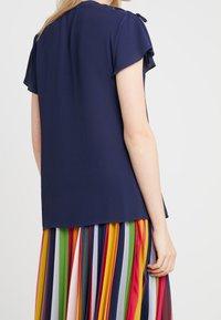 Femme portant un haut à manches courtes bleu marine avec une jupe plissée à rayures verticales multicolores, vue de dos et de côté.