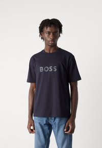 Jūrinės spalvos medvilnės marškinėliai su tekstūruotu "BOSS" logotipu šviesiai pilka spalva. Apvalus kaklas ir trumpi rankovės, derinami su mėlynomis džinsinėmis kelnėmis.