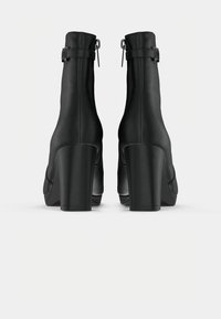 Une paire de bottines noires avec des talons hauts épais et des sangles à boucle sur le côté, présentée de dos sur un fond blanc.