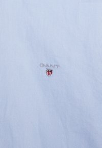 Lichtblauwe stoffen shirt met geborduurd "GANT" logo en schildontwerp. Glad textuur met een klassieke kraag en schone stikdetails.