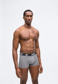 Μυώδης άντρας που φοράει γκρι μποξέρ Lacoste στέκεται μπροστά σε λευκό απλό φόντο, κοιτάζοντας μπροστά με ουδέτερο ύφος.