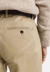 Pantalons kaki avec une poche arrière dotée d'un bouton sombre et d'une étiquette de marque. Le tissu semble lisse avec une coupe ajustée.