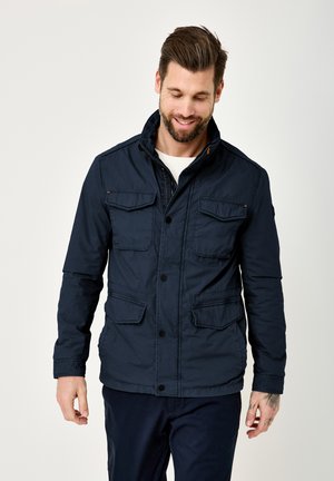 Mann mit Bart, der eine marineblaue, geknöpfte Jacke mit vier vorderen Taschen trägt, lächelnd und nach unten blickend vor einem schlichten Hintergrund.