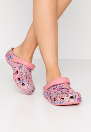 Ben iført lyserøde slip-on klipklapper med blomsterprint og justerbare hælremme på hvidt gulv og baggrund.