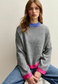 Grijze trui gemaakt van zacht materiaal, met een ronde halslijn en blauwe bies en roze ribgebreide accenten bij de mouwen en de zoom. Draag met denim.