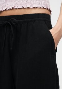 Zone de la taille avec pantalon noir à cordon élastique et main dans la poche droite, associé à un haut court rose texturé avec ourlet volanté.