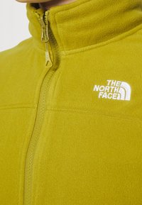 Gelbe Fleecejacke mit hohem Kragen, Frontreißverschluss und weißem Logo-Detail auf der Brust. Weiche Textur und flache Nähte sind sichtbar.