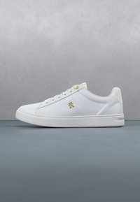 Basket basse blanche avec logo doré sur le côté, lacets blancs et une semelle blanche épaisse sur une surface grise et un fond gris clair.