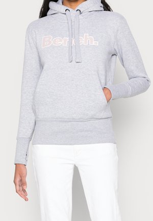 Vrouw draagt een lichtgrijze hoodie met het "Bench." logo en witte broek, hand in de zak, tegen een effen lichte achtergrond.