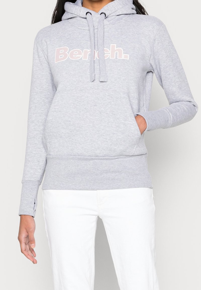 Femme portant un sweat à capuche gris clair avec le logo "Bench." et un pantalon blanc, main dans la poche, sur un fond clair uni.