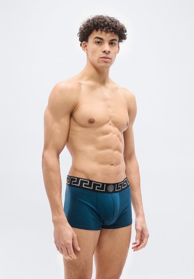 Μυώδης άντρας με σγουρά μαλλιά που φοράει πετρόλ μποξεράκια με μαύρη μέση και γεωμετρικά σχέδια με λεπτομέρεια μετάλλου.