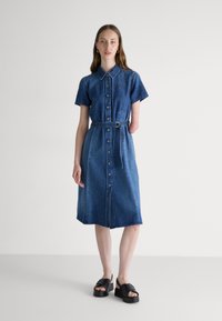 Denim shirt dress in dark blue with short sleeves, button front, and waist belt. Features a straight cut and below-knee length. Black platform sandals.
