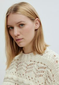 Strickpullover in Creme, mit einem offenen Spitzenmuster und geripptem Kragen. Das Design zeigt Struktur und ausgefeilte Detailarbeit.