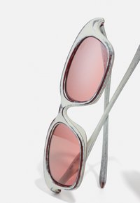Baltas vintage stila saulesbrilles ar rozā lēcām, nedaudz novecojušām malām un teksturētām pelēkām kājām, kas parāda unikālu dizaina detaļu.
