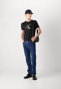 Mustvalge T-särk valge "CK" logo, sinised teksapüksid, mustad kingad ja must müts. Aksessuaaride hulka kuulub must risti keha kott.