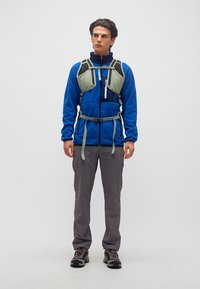 Νεαρός άνδρας που φοράει μπλε μπουφάν από φλις, γκρι παντελόνια πεζοπορίας, παπούτσια πεζοπορίας και ανοιχτοπράσινο θώρακα, στέκεται μπροστά σε ένα απλό φόντο.