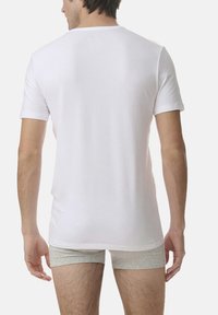 White cotton t-shirt with short sleeves, round neckline, and a smooth texture, worn over gray boxer shorts. Back view of the garment.