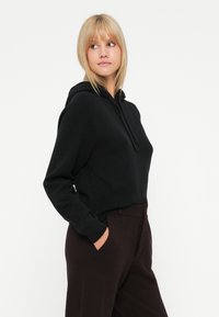 Femme blonde portant un hoodie noir et un pantalon marron foncé, les mains dans les poches, regardant légèrement vers sa gauche sur un fond blanc.
