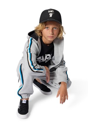 Blond child with blue eyes squats wearing grey tracksuit with blue stripes, black graphic tee, black sneakers, and black cap with white design.