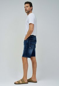 Pantalones cortos de mezclilla en azul oscuro con un acabado desgastado, combinados con una camiseta blanca lisa y sandalias de tiras marrones. Las características incluyen bolsillos frontales y detalles en la parte trasera.