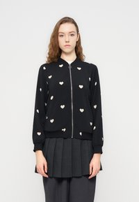 Μαύρο μπουφάν τύπου bomer με φωτεινά μπεζ σχέδια καρδιάς, φερμουάρ εμπρός, ελαστικές μανσέτες και τελείωμα. Φορεμένο πάνω από μία πλισέ γκρι φούστα.