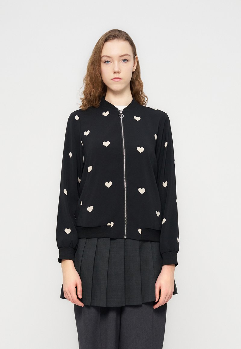 Μαύρο μπουφάν τύπου bomer με φωτεινά μπεζ σχέδια καρδιάς, φερμουάρ εμπρός, ελαστικές μανσέτες και τελείωμα. Φορεμένο πάνω από μία πλισέ γκρι φούστα.