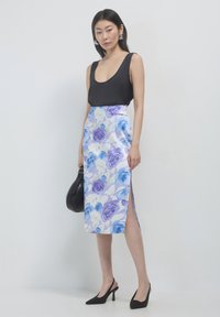 Femme debout portant un haut sans manches noir, une jupe midi blanche avec un imprimé floral bleu et violet, des talons noirs et tenant un sac à main noir.