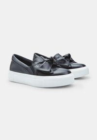 Must musta daamid mustad libised sneakers, millel on suur paeladetail, valge tekstureeritud kummitald ja sile pind.