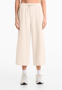 Beige wide-leg pants made of soft fabric, featuring an elastic waistband with a drawstring. Styled with white ankle socks and sneakers.