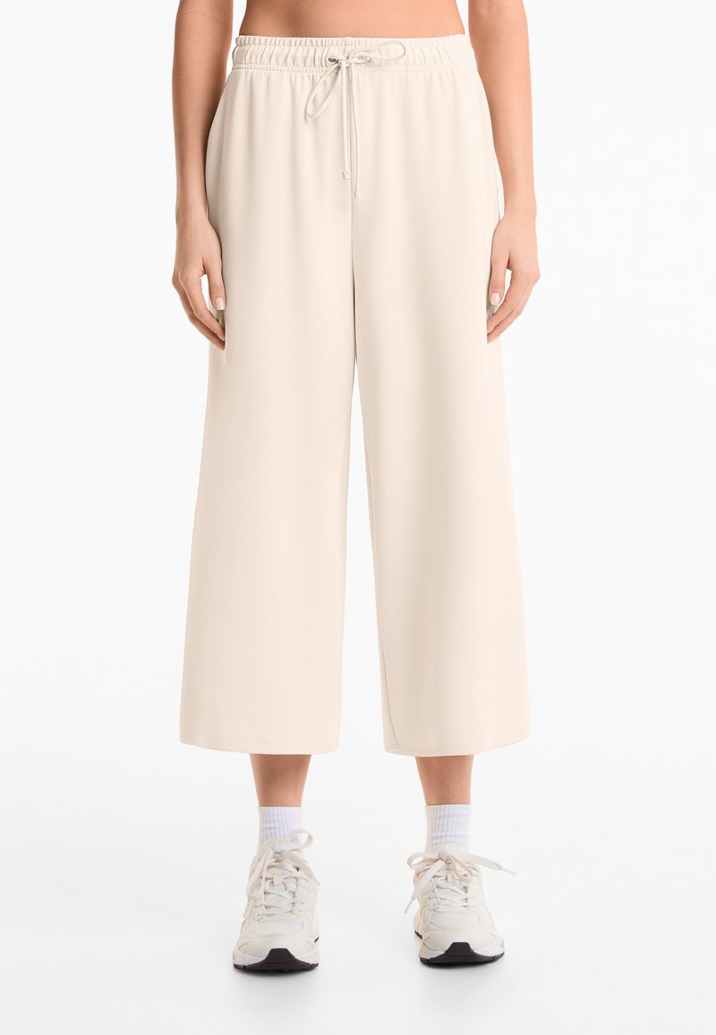 Beige weite Hosen aus weichem Stoff mit einem elastischen Taillenbund und Kordelzug. Kombiniert mit weißen Knöchelstrümpfen und Turnschuhen.
